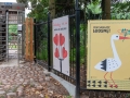 Średnie ujęcie plenerowe przedstawiające strefę wejściową do Muzeum Wsi Lubelskiej. Po lewej stronie widoczny jest nowoczesny, metalowy kołowrót (bramka obrotowa) ze skanerem biletów oraz napis „WEJŚCIE” na górnym panelu. Przed bramką ciągnie się nawierzchnia z brukowej kostki (kocich łbów), przemieszana z ziemią i opadłymi liśćmi. Prawą stronę zdjęcia zajmuje czarny, metalowy płot, na którym zamontowane są dwa pionowe plakaty z wystawy prac Łukasza Tarkowskiego: Plakat po prawej (Pierwszy plan): Żółty plakat w drobne, zielone kreskowanie tworzące ramkę. Przedstawia dużą, stylizowaną grafikę białego bociana z czerwonym dziobem i nogami, który trzyma w dziobie gałązkę z czerwonymi owocami/kwiatami. W lewym górnym rogu znajduje się czarny napis: „PRZYWOŁAJMY WIOSNĘ!”. U dołu plakatu widnieje rząd małych, czarnych sylwetek ptaków. Plakat po lewej (Bliżej bramki): Jasnoszary/biały plakat z czerwowym, pisanym odręcznie napisem u góry: „Zakochaj się w” oraz drukowanym poniżej: „MUZEUM WSI LUBELSKIEJ”. Centralną grafikę stanowią czarne widły, na których ułożone są cztery czerwone serca. Obraz ten można również interpretować jako czarną gałąź/drzewo, na której ułożone są cztery czerwone serca imitujące liście lub owoce. Za płotem i w tle widać bujną zieleń drzew i krzewów skansenu. Światło jest naturalne i rozproszone.